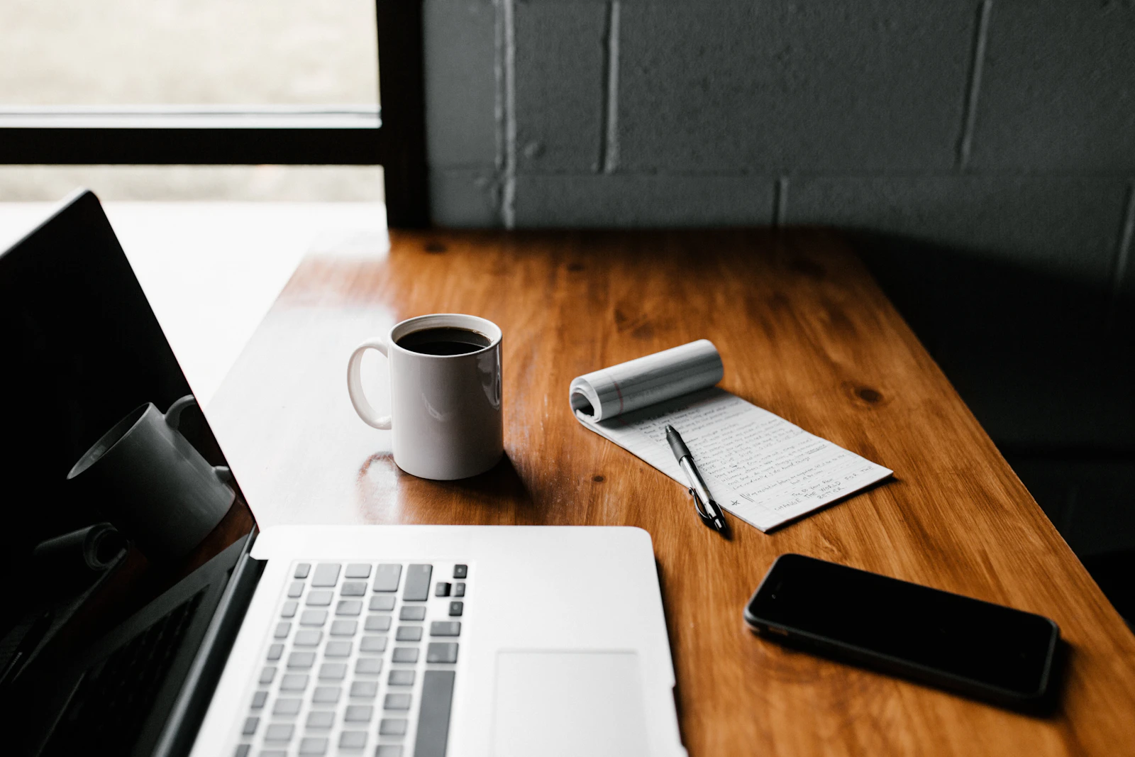 A laptop, notebook, coffee, and pen arranged on a wooden workspace for planning digital content